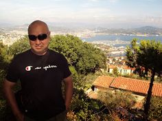 a man wearing sunglasses standing in front of some trees and buildings with the ocean in the background