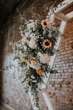 flowers and greenery are arranged on the side of a wooden ladder in front of a brick wall