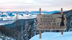 a wooden sign on top of a snow covered slope with mountains in the background and trees