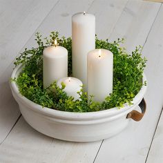 three white candles sitting in a bowl filled with greenery