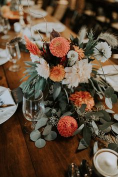 an arrangement of flowers and greenery on a wooden table at a wedding reception with place settings