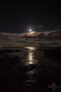 the moon is shining in the night sky over the water and rocks on the shore