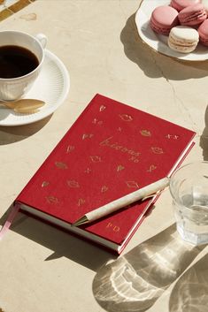 a red notebook sitting on top of a table next to a cup of coffee and macaroons