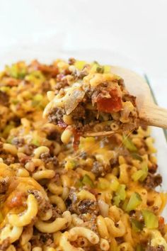 a wooden spoon full of macaroni and cheese with meat on the top, in a glass casserole dish