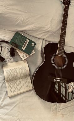 an acoustic guitar, headphones and book laying on a bed with the sheets pulled back