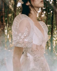 a woman in a white dress standing in the woods with her hair up and flowers in her hair