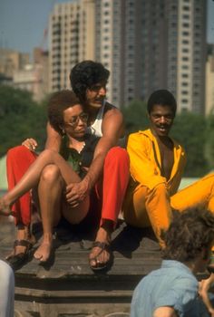 three men sitting on the back of a truck in front of some buildings and people