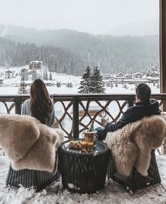 two people sitting on chairs in front of a balcony with snow covered mountains and trees