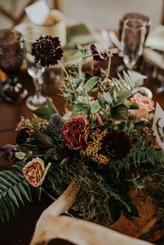 an arrangement of flowers and greenery is displayed on a table with wine glasses, napkins and place cards