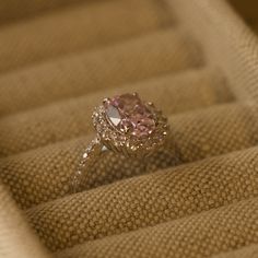 a pink diamond ring sitting on top of a tan cloth covered box with gold trim