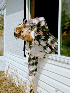 a woman leaning on the side of a house with her hands in her hair and wearing floral pants