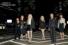 four women standing in the middle of an empty parking lot at night with skyscrapers in the background