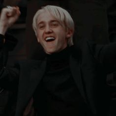 a man with white hair is smiling and raising his arms in the air while wearing a black suit