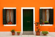 an orange building with black shutters and two green doors on each side, surrounded by potted plants