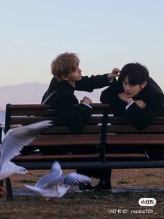 two young boys sitting on a bench with seagulls flying around them in front of them