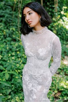 a woman wearing a white dress standing in front of some bushes and trees with her hands on her hips