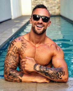 a man with tattoos and sunglasses sitting in front of a swimming pool smiling at the camera