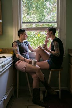 two people sitting at a kitchen window sill eating food and drinking from a cup