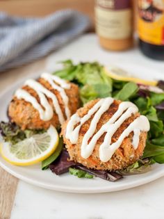 two crab cakes on a white plate with greens and lemon wedges next to them