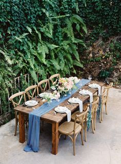 an outdoor table set up with plates and place settings for six people, surrounded by greenery