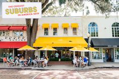 people sitting at tables under yellow umbrellas in front of a building with red and white awnings