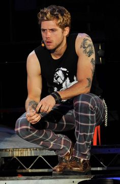 a man sitting on top of a skateboard while wearing plaid pants and a black tank top