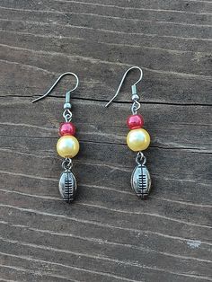 two pairs of earrings with football charms hanging from them on a wooden surface, one is yellow and the other is red