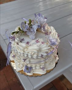 a white cake with purple flowers on top sitting on a picnic table in the sun