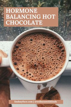 a person holding a cup of hot chocolate in front of a window with rain drops on it