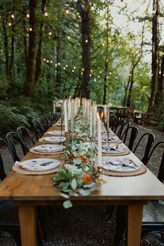 a long table with candles and greenery is set for an outdoor dinner in the woods