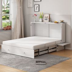 a white bed with drawers underneath it in a living room next to a large window