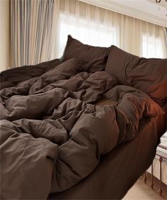 an unmade bed with brown sheets and pillows in front of a curtained window