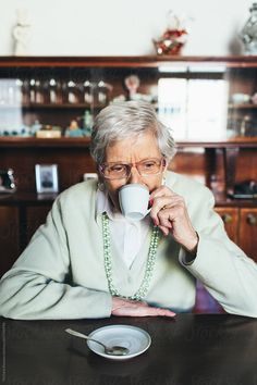 an elderly woman drinking from a coffee cup while sitting at a table in front of a bookcase