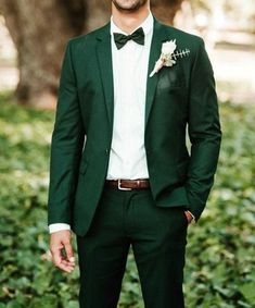 a man wearing a green suit and bow tie standing in front of some bushes with his hands in his pockets