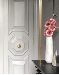 a white vase with pink flowers sitting on a black table in front of a door