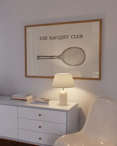 a white chair sitting next to a table with a lamp on it and a racket poster above it