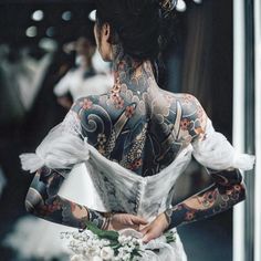 the back of a woman's body with tattoos on her arms and shoulder, holding a bouquet of flowers