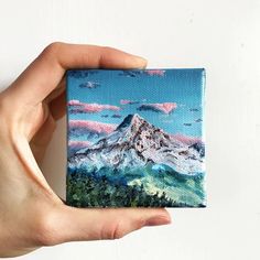 a hand holding up a small painting with mountains in the background