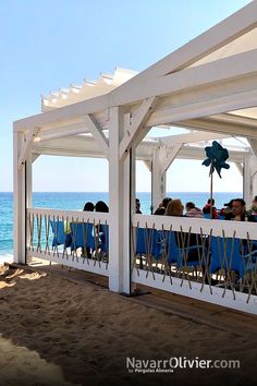 some people are sitting on chairs under a white gazebo by the beach with blue water