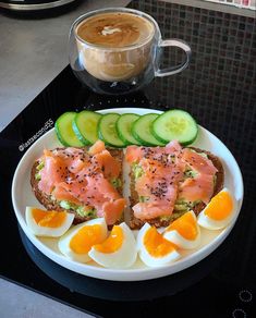 a plate with eggs, salmon and cucumbers on it next to a cup of coffee