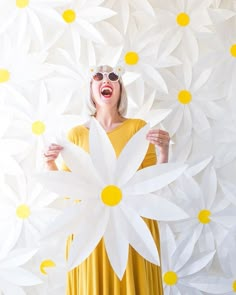 a woman in yellow dress holding up large white paper flowers with sunglasses on her head