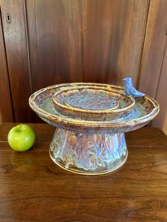 a blue bird sitting on top of a glass dish next to an apple and wood paneled wall