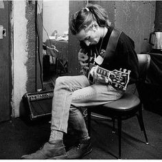 a man sitting in a chair holding a guitar and looking down at his cell phone