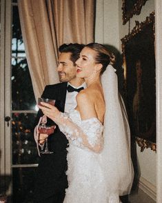 a bride and groom standing next to each other in front of a window holding wine glasses