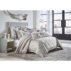 a bed with white and silver comforters in a bedroom next to a window overlooking the city