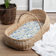 an empty wicker basket next to a plant and towel on a wooden floor with a potted plant in the background