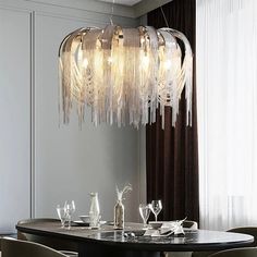 A modern dining room showcasing a Morsale.com Cadena Contemporary Chandelier, featuring adjustable hanging wire and cascading metallic strands with LED bulbs illuminating a dark oval table set with elegant glassware and minimalistic décor. The backdrop is adorned with floor-to-ceiling curtains and a light grey wall. Sculptural Glass Chandelier Dining Room, Crystal Dripping Chandelier Low Clearance, Modern Chandelier Dining, Smoked Glass Chandelier, Modern Chrystal Chandelier, Contemporary Chandeliers, Dining Room Chandelier Modern, Dining Chandelier, Christal Chandelier Murano