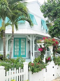 a white house with blue shutters and flowers on the front porch, next to a palm tree