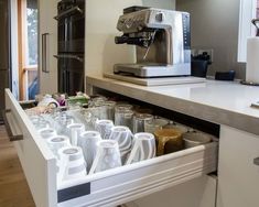 an open drawer in a kitchen filled with glasses and coffee maker sitting on top of a counter