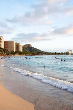 many people are swimming in the ocean on a sunny day at the beach with tall buildings behind them